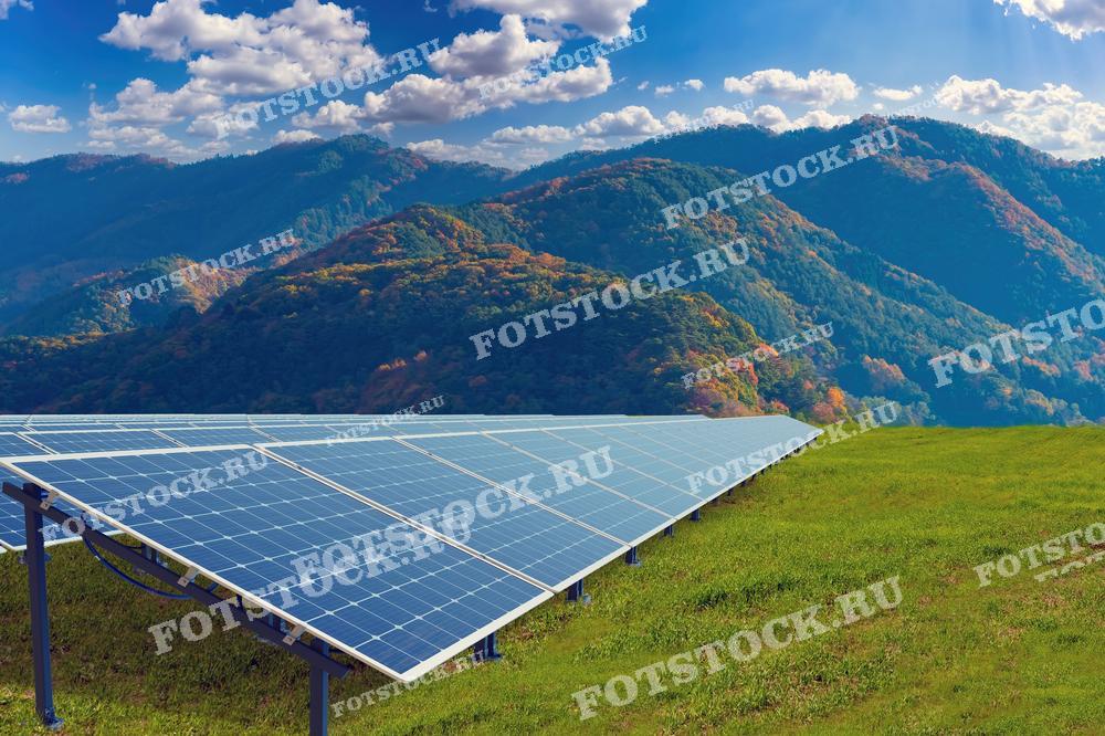 Фотография изображает солнечные панели, установленные на фоне живописных гор и зеленой травы под голубым небом с облаками. Этот снимок подчеркивает гармоничное сочетание возобновляемой энергии и природной среды.